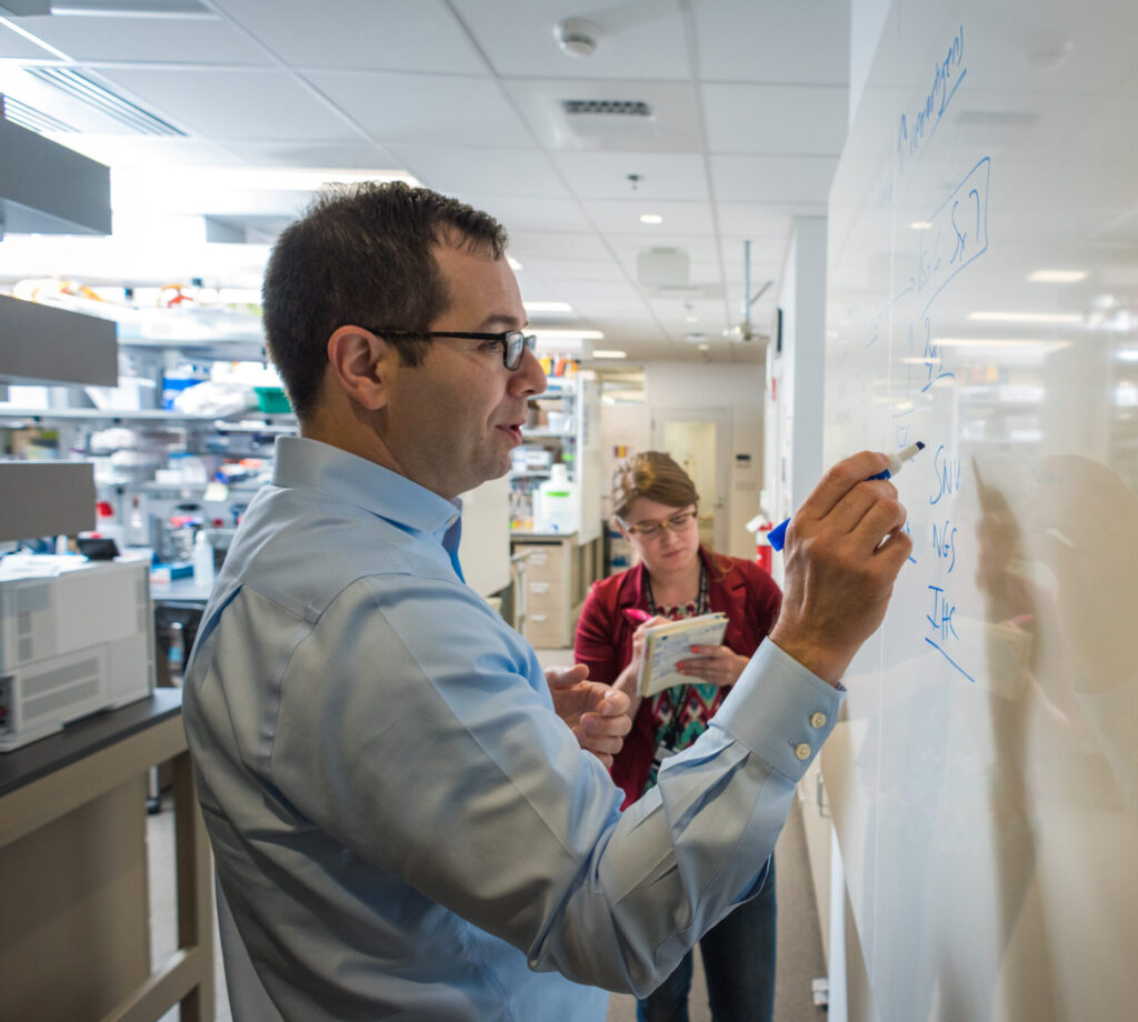 Sandro Santagata and lab member discuss research insights inside LSP lab space. Santagata writes on white board inside LSP lab while team member takes notes in background.