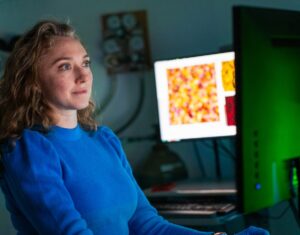 Woman in blue looking at computer screen. Another screen in the background displays a yellow and red image. 